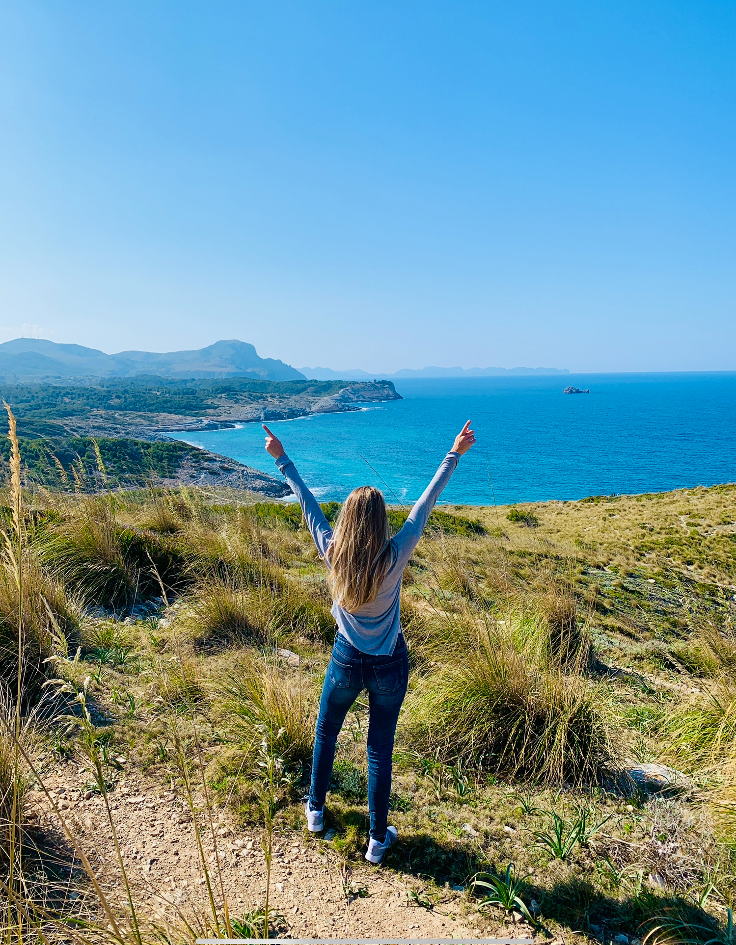 Junge Frau mit erhobenen Armen an einem Aussichtspunkt im Nordosten Mallorcas, umgeben von Küstenschilf, mit Blick auf das türkise Meer und Kap Formentor im Hintergrund.
