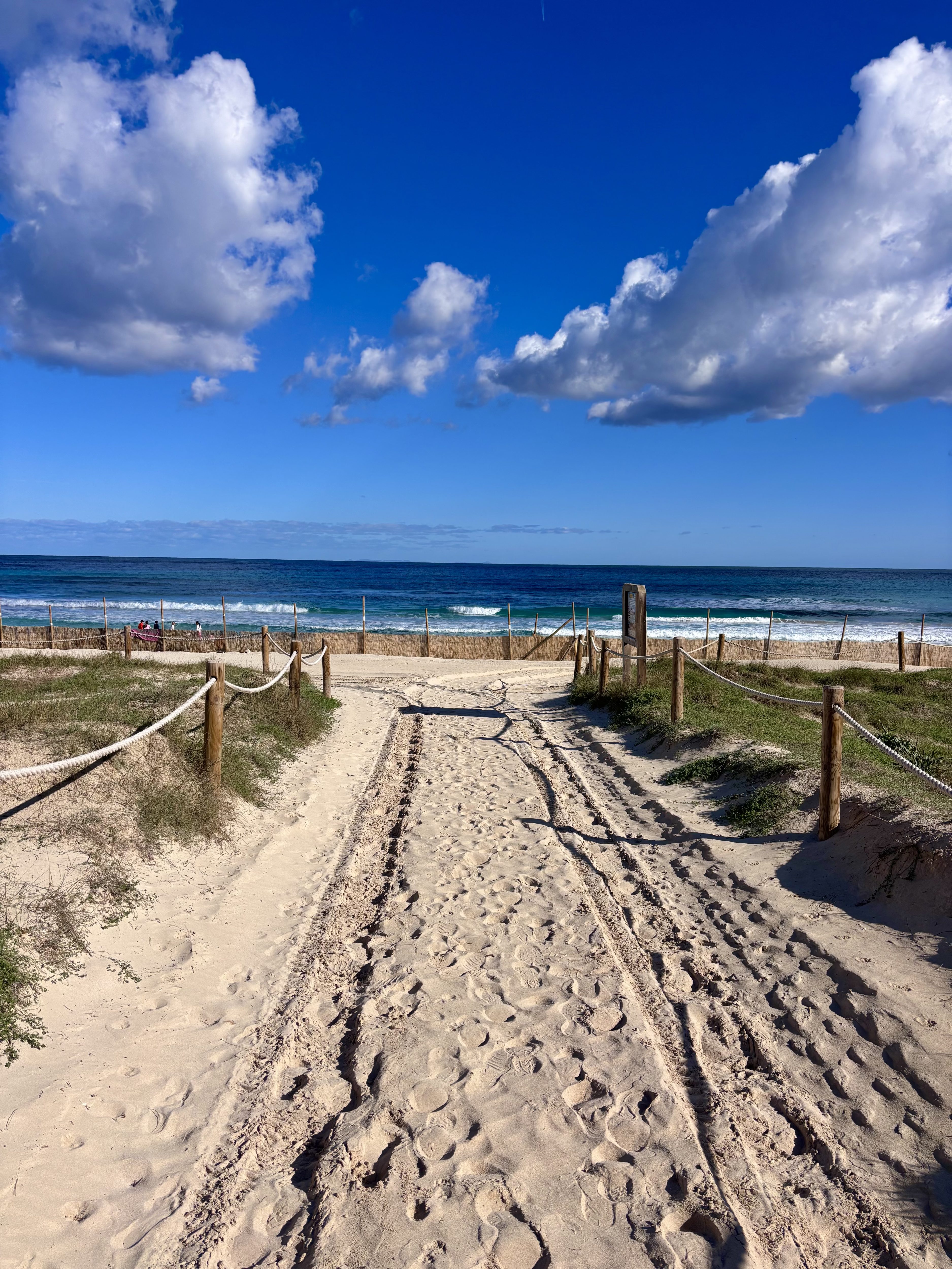 Strandzugang zur Cala Agulla mit seitlichen Holzpfosten und Seilen, Blick über den sandigen Weg direkt auf das blaue Meer unter sonnigem Himmel mit ein paar Wolken.